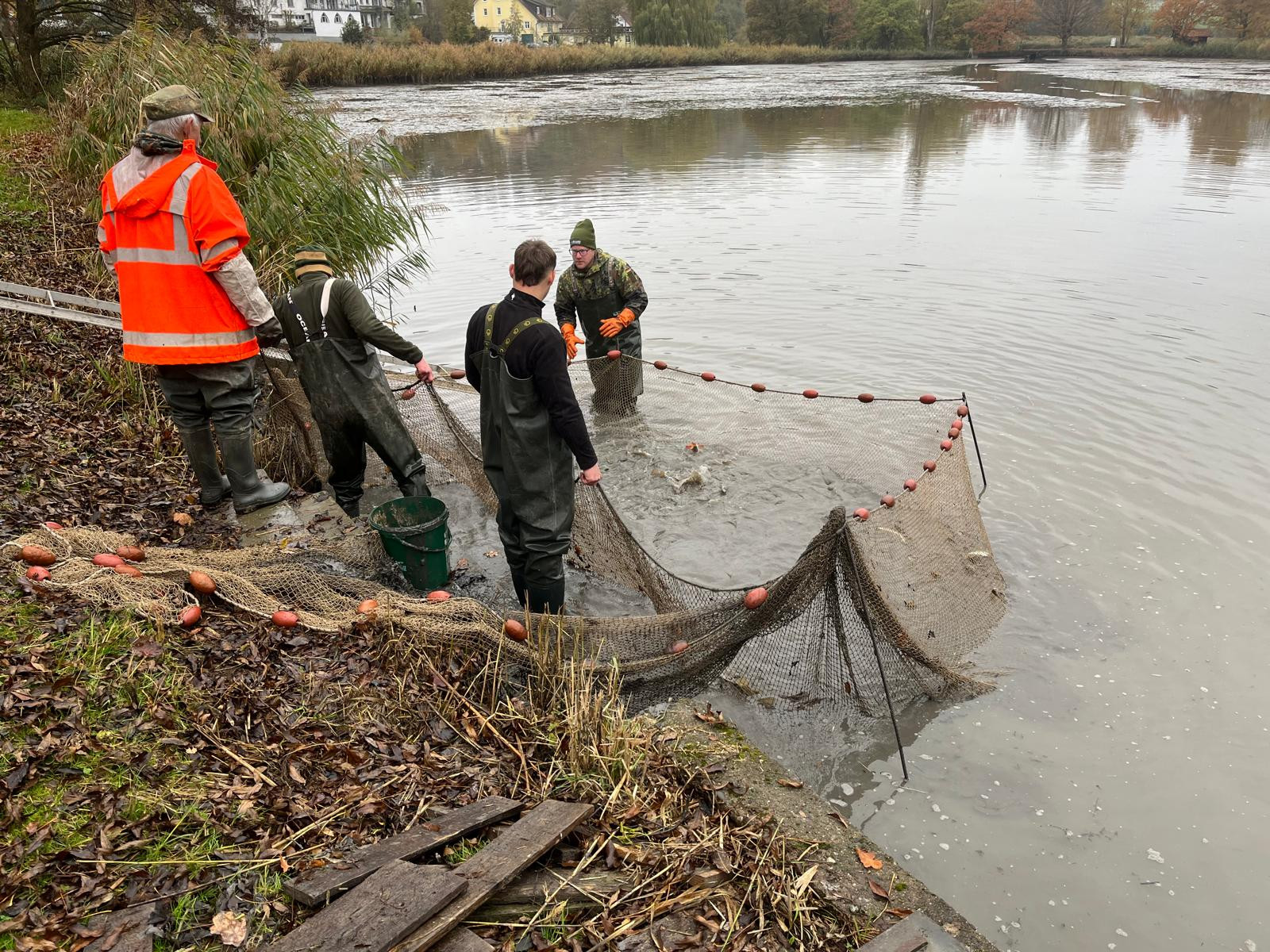 KI generiert: Auf dem Bild sind mehrere Personen zu sehen, die in einem Gewässer ein Netz auslegen, offenbar um Fische zu fangen. Sie stehen teils im Wasser und tragen wetterfeste Kleidung.